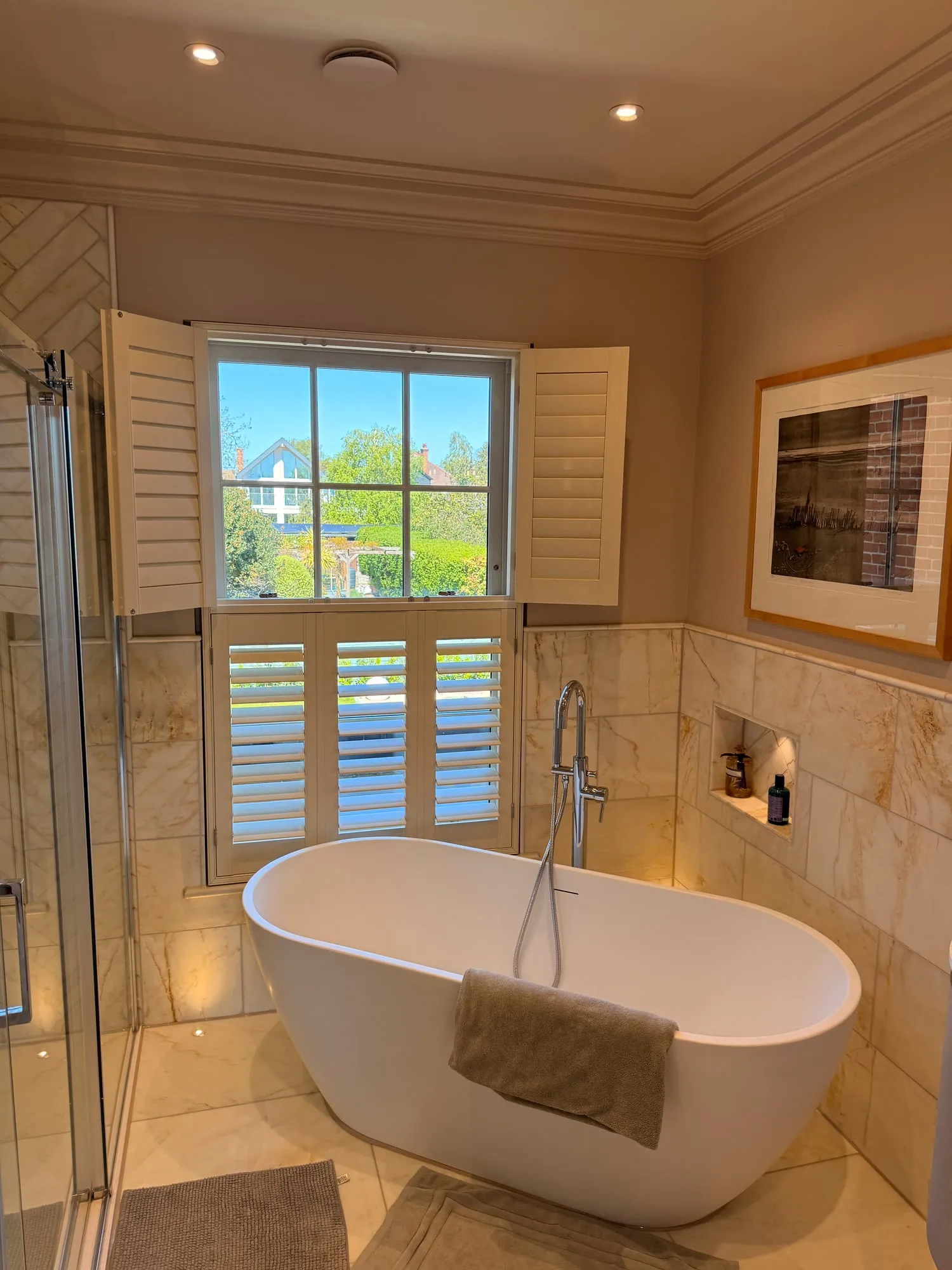 Café-style shutters above a freestanding oval bath in a marble Suffolk bathroom