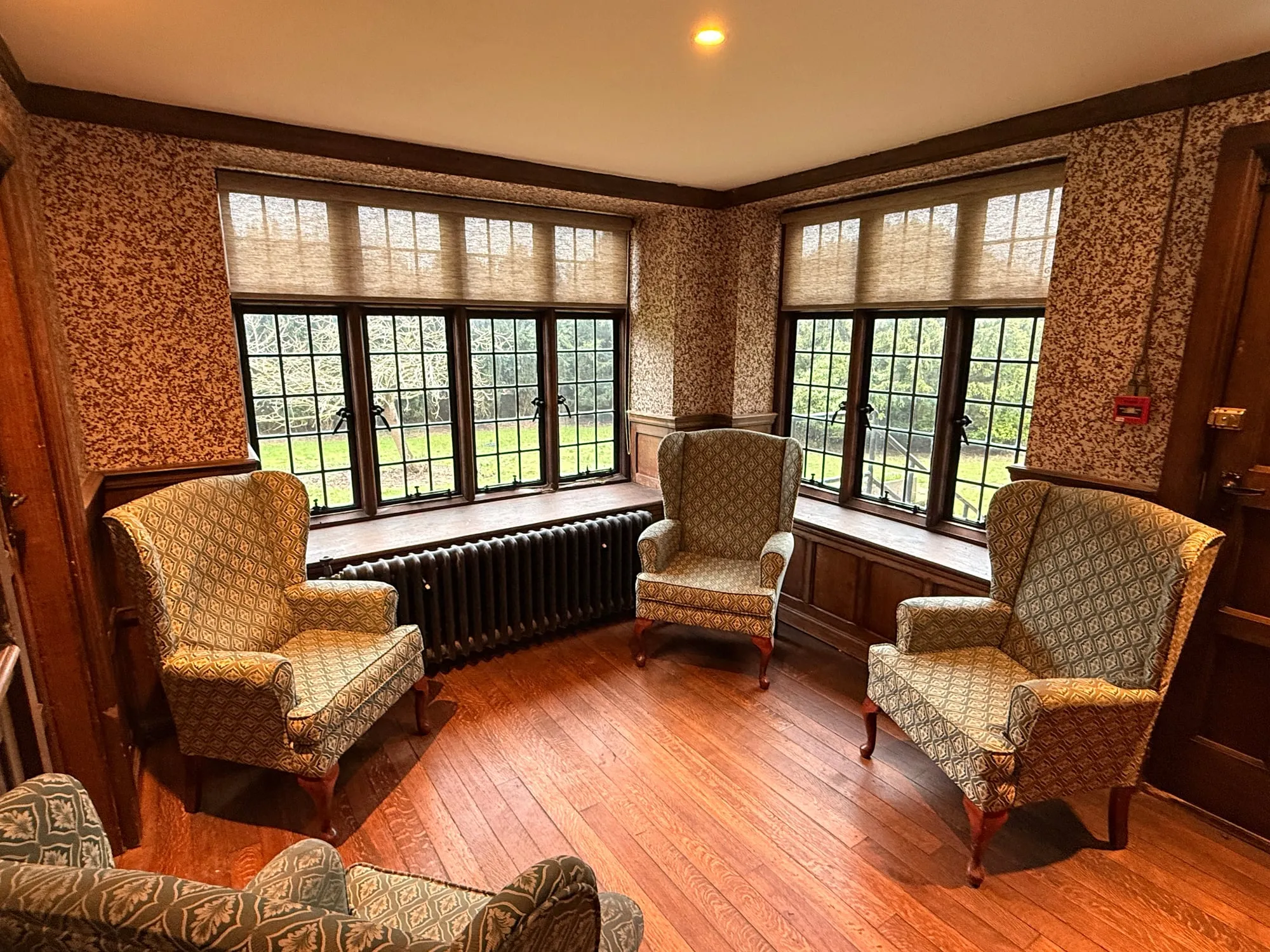 Textured cream roller blinds on a leaded bay window in a Suffolk period sitting room with oak panelling and wing chairs